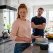 Woman surprised by smart speaker playing a voice note in a kitchen, man watching silently