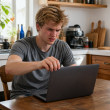 Young man sitting at kitchen table opening laptop, frustrated by Wi-Fi drop