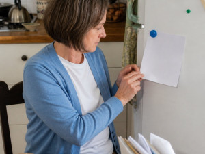 Woman sitting at kitchen table looking at 'No Yelling' rule on fridge, hands fidgeting, kitchen background.