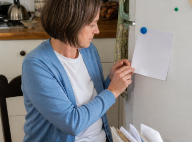 Woman sitting at kitchen table looking at 'No Yelling' rule on fridge, hands fidgeting, kitchen background.