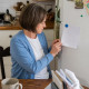 Woman sitting at kitchen table looking at 'No Yelling' rule on fridge, hands fidgeting, kitchen background.