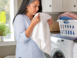 Latina woman sniffing a towel in laundry room, puzzled expression, casual clothes.