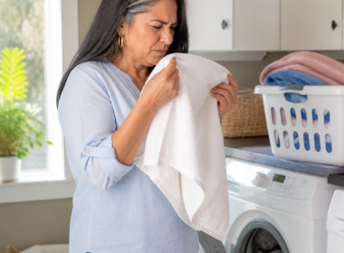 Latina woman sniffing a towel in laundry room, puzzled expression, casual clothes.