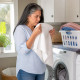 Latina woman sniffing a towel in laundry room, puzzled expression, casual clothes.