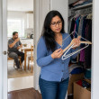 Woman looking at empty hangers in closet while man drinks coffee at table nearby.