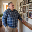 Man in blue flannel shirt examining a framed family photo wall in a cozy hallway.