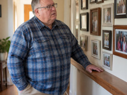 Man in blue flannel shirt examining a framed family photo wall in a cozy hallway.