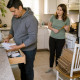 Evan sorts manuals and receipts at the kitchen counter while Mara watches from the stove, spoon paused.