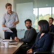 In a conference room, Dylan gestures while looking too closely at Mara as coworkers quietly notice.