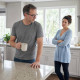 Grant shields a face-down phone on the kitchen counter while Nina watches him closely.