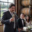 A man mid-toast freezes as he stares at a place card, with the smiling groom and tense bride behind him.