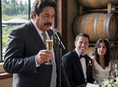 A man mid-toast freezes as he stares at a place card, with the smiling groom and tense bride behind him.