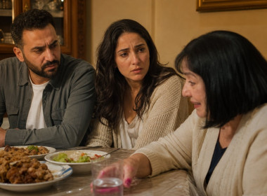 At a tense family dinner, the older woman reaches for a water glass with visibly stained fingers while the couple watches.