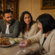 At a tense family dinner, the older woman reaches for a water glass with visibly stained fingers while the couple watches.