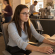 A tense young woman with glasses at her office desk, frozen mid-reach as her phone vibrates beside her.
