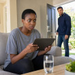 Mara stares at a tablet on the couch as Evan walks in through the front door.