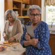 A shocked older Black woman clutches a tablet in a kitchen while her mother calmly slices peaches behind her.