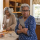 A shocked older Black woman clutches a tablet in a kitchen while her mother calmly slices peaches behind her.