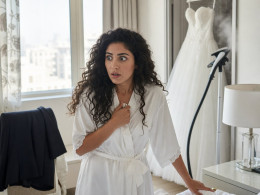 Bride in a satin robe clutches a phone, stunned, beside an unmade bed with a suit jacket nearby.