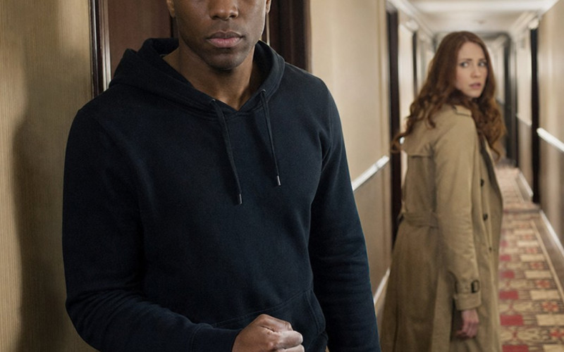 A tense young man stands by a hotel room door as a woman rounds the corner and freezes.