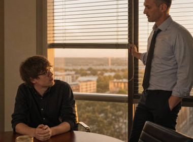 After-hours in a glass-walled conference room, Graham lowers the blinds while Iris looks up from the table, tense.