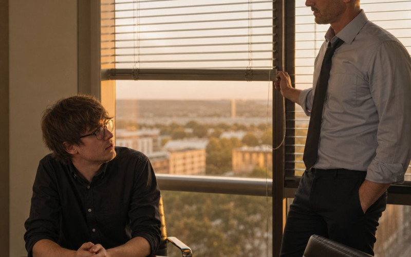 After-hours in a glass-walled conference room, Graham lowers the blinds while Iris looks up from the table, tense.