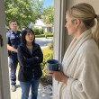 A stunned woman stands with a police officer at her door as a blonde woman in a bathrobe holds a mug and blocks the entry.