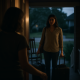 Woman opening front door to another woman with suitcases at dusk.
