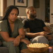 A tense woman with long box braids sits beside her relaxed husband on a couch, gripping a face-down phone and staring at him.