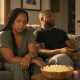 A tense woman with long box braids sits beside her relaxed husband on a couch, gripping a face-down phone and staring at him.