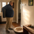 A heavyset gray-haired man with glasses stares at an empty leash hook beside a flattened dog bed.