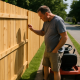 Man with lawn mower stopped near a new wooden fence cutting into grass strip beside sidewalk.