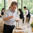 An older man with gray hair and reading glasses finds a second place card tucked beneath another at a wedding table.
