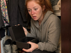 A red-ponytailed woman crouches in a closet holding an open lockbox, shocked.