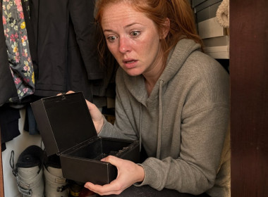 A red-ponytailed woman crouches in a closet holding an open lockbox, shocked.
