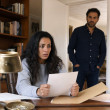 Claire grips a deed-like paper at her desk as Grant watches from the doorway, tension visible in their faces.