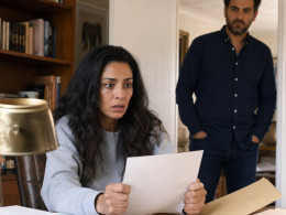 Claire grips a deed-like paper at her desk as Grant watches from the doorway, tension visible in their faces.