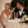 The bride grips a face-down phone under the sweetheart table while the groom turns toward her, smiling uncertainly.