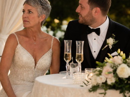 The bride grips a face-down phone under the sweetheart table while the groom turns toward her, smiling uncertainly.