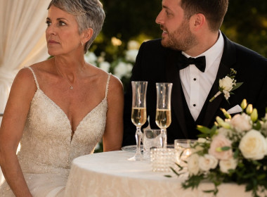 The bride grips a face-down phone under the sweetheart table while the groom turns toward her, smiling uncertainly.