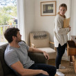 Tessa stands at Miles’s open desk drawer holding an envelope while Miles watches from the couch.