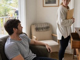 Tessa stands at Miles’s open desk drawer holding an envelope while Miles watches from the couch.