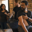 In a tattoo studio, a bearded South Asian man covers his ribs while a tattoo artist gestures toward a drawer and a woman watches in shock.
