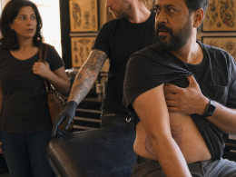 In a tattoo studio, a bearded South Asian man covers his ribs while a tattoo artist gestures toward a drawer and a woman watches in shock.