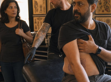 In a tattoo studio, a bearded South Asian man covers his ribs while a tattoo artist gestures toward a drawer and a woman watches in shock.