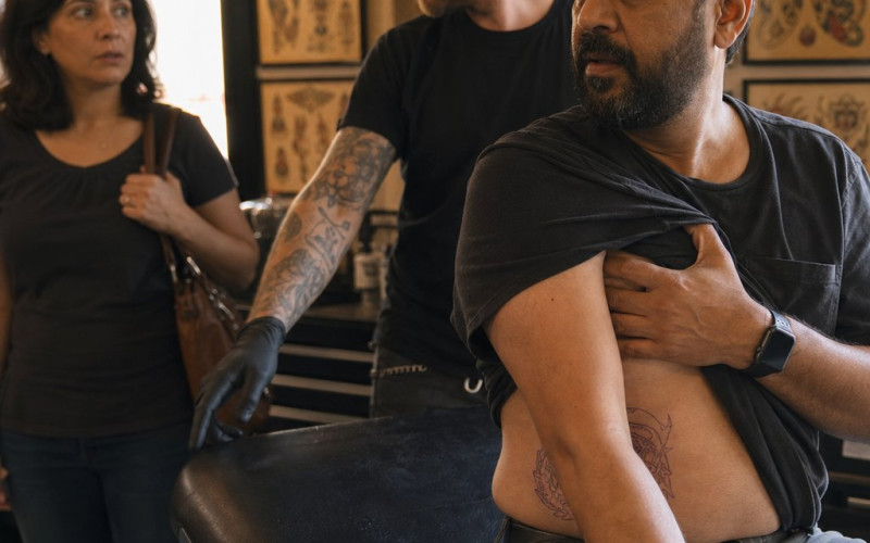 In a tattoo studio, a bearded South Asian man covers his ribs while a tattoo artist gestures toward a drawer and a woman watches in shock.