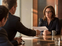 An investigator slides a printed page toward a tense woman in glasses at a boardroom table.