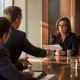 An investigator slides a printed page toward a tense woman in glasses at a boardroom table.