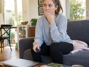 Lena sits rigid on a couch, hand to her mouth, staring ahead as if shocked by what she just saw.