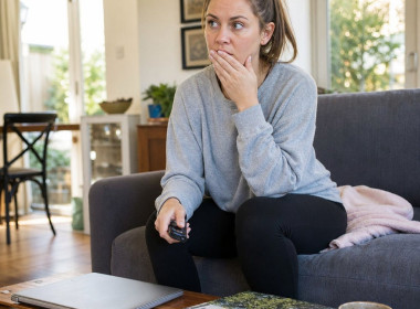 Lena sits rigid on a couch, hand to her mouth, staring ahead as if shocked by what she just saw.
